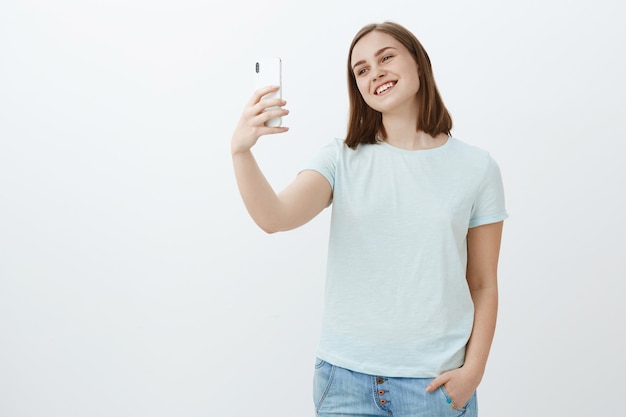 süßes glückliches und selbstbewusstes Mädchen, das mit Mutter über Videobotschaften spricht, während sie im Ausland studiert, das den Kopf des Smartphones kippt und auf dem Gerätebildschirm lächelt und Selfie über weiße Wand nimmt