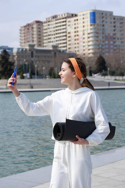 Süßes junges Mädchen, das eine gerollte Yogamatte hält und ein Selfie in hoher Qualität macht