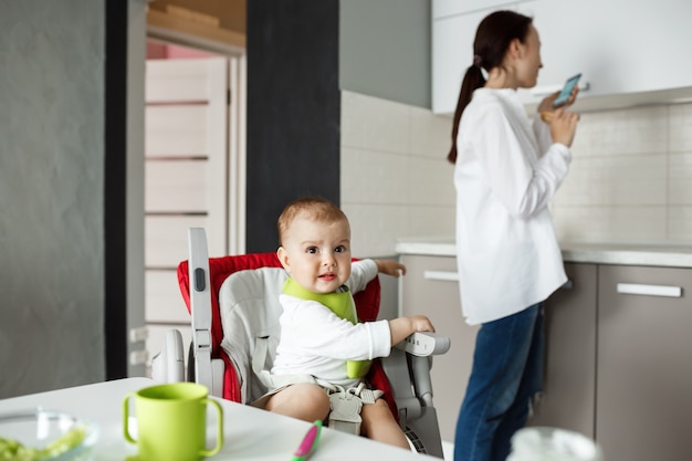 Süßes kleines Baby im grünen Lätzchen, das im Babystuhl sitzt und Angst hat, Vogel außerhalb des Fensters zu sehen, während Mutter sich zur Seite dreht, um Chatrooms auf Smartphone zu überprüfen.