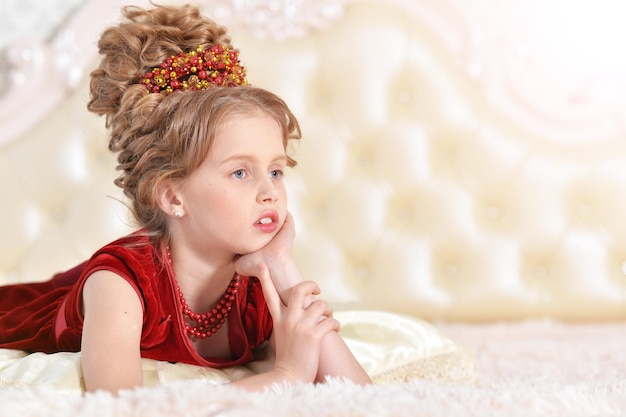 Süßes kleines mädchen im roten samtkleid mit retro-frisur auf beige couch liegend
