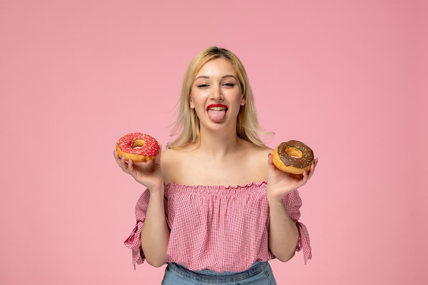 Süßes Mädchen, entzückendes blondes Küken mit roter Lippenstift-Rosa-Bluse mit herausgestreckter Zunge, die Donuts hält