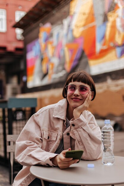 Süßes Mädchen mit Kurzhaarfrisur lächelt draußen Modische brünette Frau mit Sonnenbrille und beigem Outfit, die Telefon hält und mit Wasser im Café sitzt
