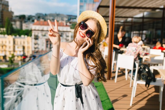 Süßes Mädchen mit langen Haaren in Sonnenbrille steht auf der Terrasse. Sie trägt ein weißes Kleid mit nackten Schultern, roten Lippenstift und Hut. Sie spricht am Telefon und lächelt in die Kamera.