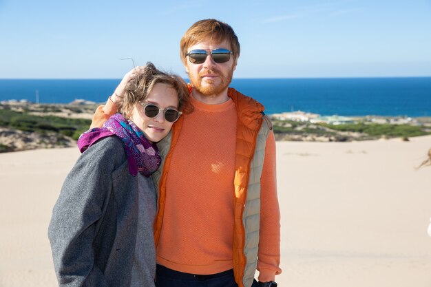 Süßes Paar in warmer Kleidung, die Freizeit auf See verbringt, auf Sand steht, umarmt, nach vorne schaut