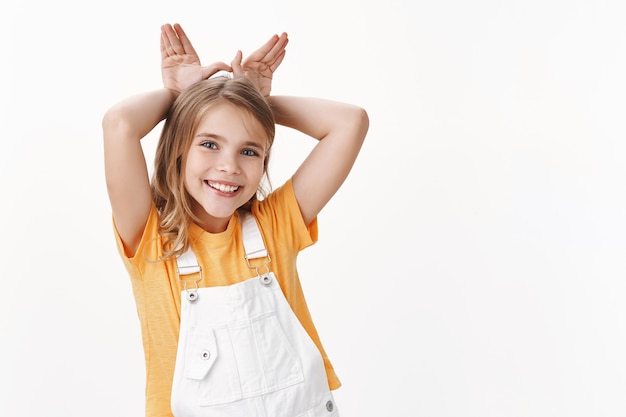 Süßes reizendes kleines Mädchen, blondes Kind im T-Shirt, Overall, Hasenohren imitieren Kaninchen, halten Handflächen hinter dem Kopf, lächeln freudig, spielen süß und zart, weiße Wand