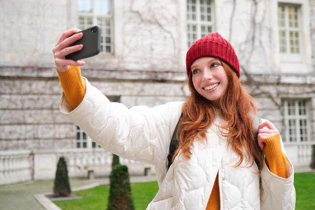 Süßes rothaariges Mädchen mit rotem Hut macht Selfie während ihrer Touristenreise ins Ausland. Junge rothaarige Frau macht a