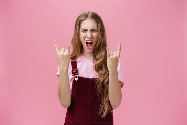 Süßes und aufgeregtes Party-Girl mit welliger Frisur in Cord-Overalls, das ja schreit und Rock-n-Roll-Geste zeigt, die Spaß daran haben, harte Musik zu genießen, die gewagt und rebellisch über rosa Wand ist. Platz kopieren
