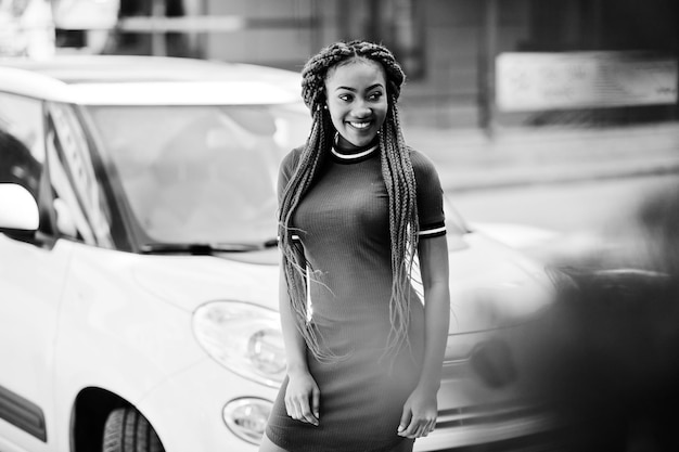 Süßes und schlankes afrikanisch-amerikanisches Mädchen in rotem Kleid mit Dreadlocks posierte im Freien auf der Straße Hintergrund kleines weißes Auto Stilvolles schwarzes Modell