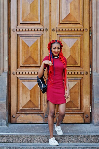 Süßes und schlankes afrikanisch-amerikanisches Mädchen in rotem Kleid mit Dreadlocks und Rucksack posierte vor der großen Holztür der Schule Stilvoller schwarzer Student
