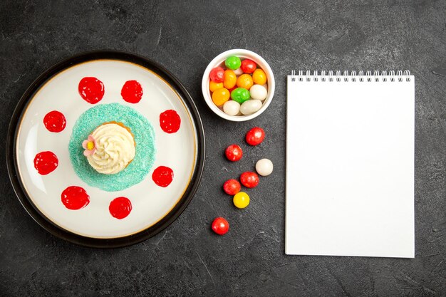 Süßigkeiten von oben auf dem Tisch appetitlicher Cupcake neben der Schüssel mit Süßigkeiten und weißem Notizbuch auf dem schwarzen Tisch