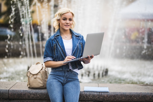 Sunny blonde studentin arbeitet an ihrem laptop in der nähe von brunnen in der stadt am tag