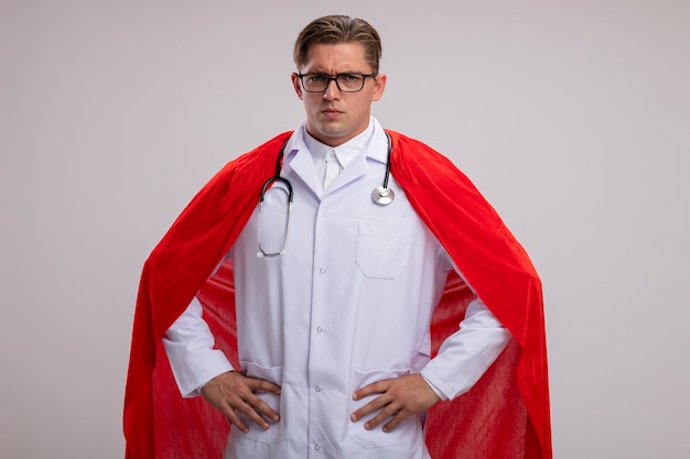 Superheld Doktormann, der weißen Mantel im roten Umhang und in den Gläsern mit Stethoskop um den Hals trägt, betrachtet Kamera mit ernstem sicherem Ausdruck mit Armen an der Hüfte, die über weißem Hintergrund stehen