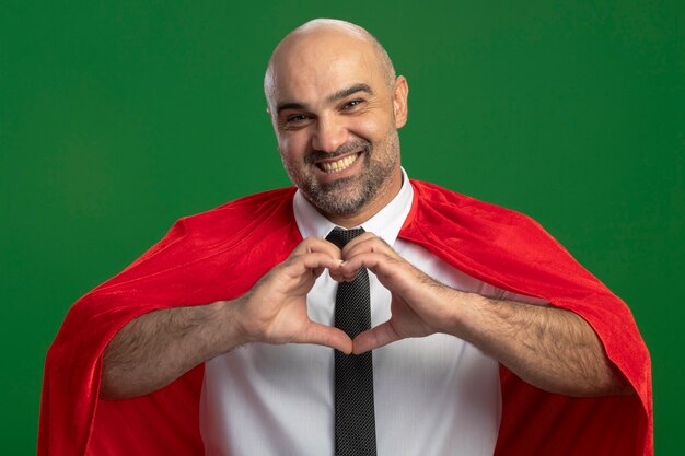 Superheld-Geschäftsmann im roten Umhang, der Herzgeste mit den Fingern macht, die vorne lächelnd fröhlich über grüner Wand stehend schauen