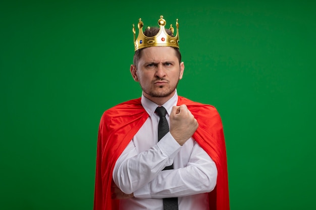 Superheld-Geschäftsmann im roten Umhang, der Krone mit ernstem Gesicht trägt, das über grüner Wand unzufrieden steht