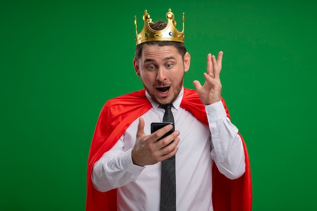 Superheld-Geschäftsmann im roten Umhang, der Krone unter Verwendung des Smartphones trägt, das erstaunt und überrascht über grünem Hintergrund steht