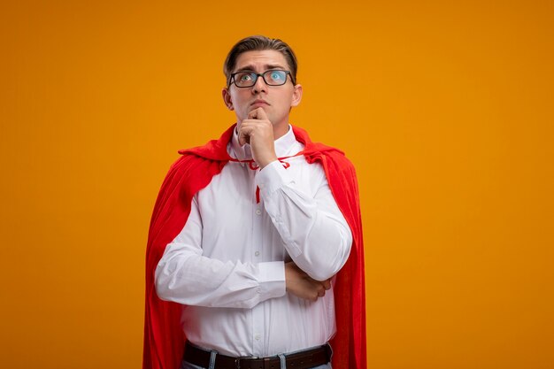 Superheld-Geschäftsmann im roten Umhang und in den Gläsern, die beiseite schauen, verwirrt über orange Hintergrund