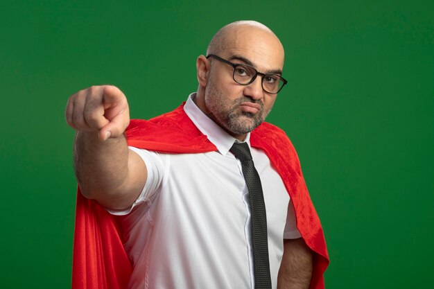 Superheld-Geschäftsmann im roten Umhang und in den Gläsern, die mit Zeigefinger auf Kamera zeigen, die über grüner Wand unzufrieden steht
