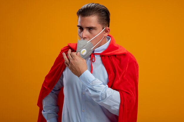 Superheld-Geschäftsmann in der schützenden Gesichtsmaske und im roten Umhang, die müde und gelangweilt aussehen und Maske abnehmen, die über orange Wand steht