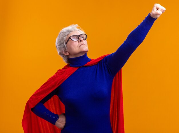 Superheldin der älteren Frau mit Brille mit rotem Umhang, die nach oben schaut und Gewinnergeste mit der Hand macht, die bereit ist, zu helfen und zu kämpfen