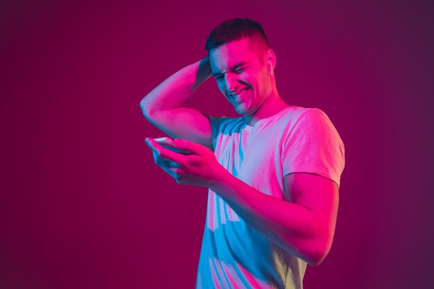 Surfen, Wetten, Zuschauen, Selfie. Porträt des kaukasischen Mannes isoliert auf rosa-violetter Wand im Neonlicht. Männliches Model mit Geräten. Konzept der menschlichen Emotionen, Gesichtsausdruck,