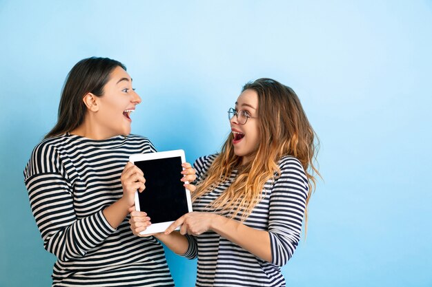 Tablet mit leerem Bildschirm halten. Junge emotionale Frauen lokalisiert auf blauem Studiohintergrund der Steigung. Konzept der menschlichen Emotionen, Gesichtsausdruck, Freundschaft, Anzeige. Schöne Modelle in Freizeitkleidung.