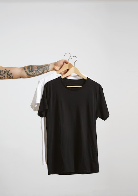 Tätowierte Bikerhand hält Holzhänge mit leeren schwarzen und weißen T-Shirts aus hochwertiger dünner Baumwolle, isoliert auf Weiß
