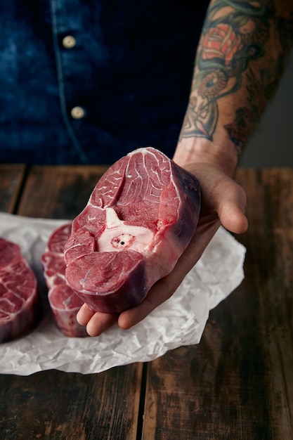 Tätowierte Hand bietet ein Stück Fleischpfahl über zwei Steaks auf Bastelpapier, Nahaufnahme