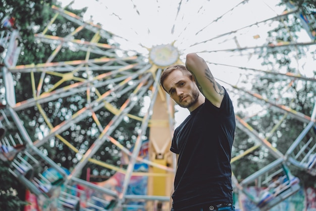 Kostenloses Foto tätowierter junger mann in einem vergnügungspark auf dem hintergrund eines karussells