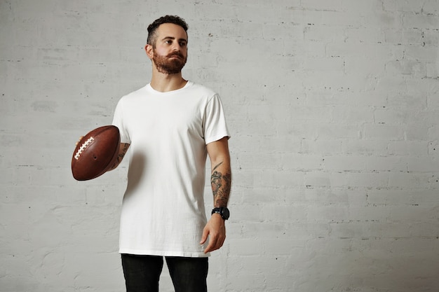 Tätowiertes und bärtiges Modell im schlichten weißen Kurzarm-T-Shirt, das einen Lederfußball auf grauer Wand hält