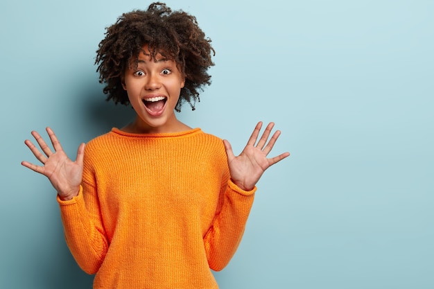 Kostenloses Foto taille hoch aufnahme von fröhlichen dunkelhäutigen frau zeigt handflächen, ruft vor freude, reagiert auf schönes geschenk, hat glücklichen gesichtsausdruck, trägt orangefarbenen pullover, isoliert über der blauen wand. emotionskonzept