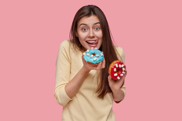 Taille hoch Schuss der glücklichen kaukasischen Dame leckt Lippen mit der Zunge, hält köstliche Donuts, hält sich nicht an Diät