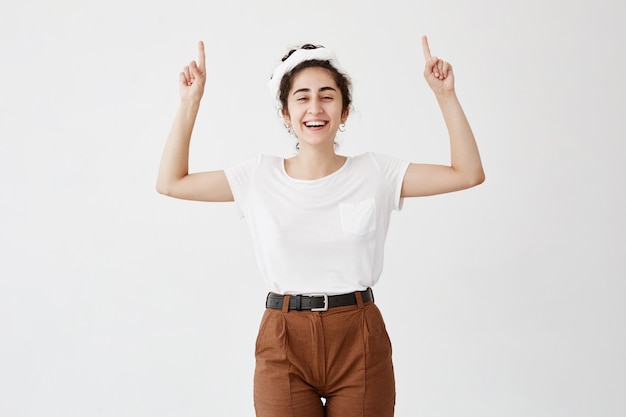 Taille hoch Schuss des freudigen glücklichen Mädchens, das weißes T-Shirt trägt und Finger auf Kopierraum über ihrem Kopf zeigt. Junge Frau, die etwas auf leere Wand mit Händen anzeigt