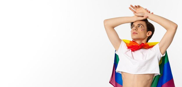 Taillenaufnahme eines sinnlichen androgynen Mannes mit bauchfreiem Oberteil und Regenbogen-Stolzflagge, der mit erhobenen Händen auf den weißen Hintergrund auf die obere linke Ecke blickt