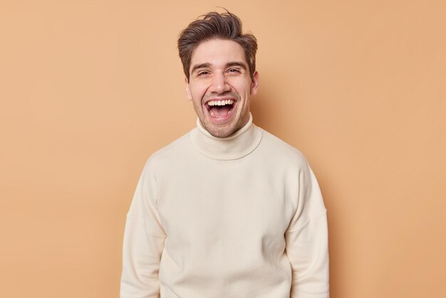 Taillenaufnahme eines überglücklichen gutaussehenden Mannes, der glücklich lacht und aufrichtige Gefühle ausdrückt, hört etwas Lustiges, das in einem lässigen weißen Pullover gekleidet ist, der über braunem Hintergrund isoliert ist Menschen- und Glückskonzept