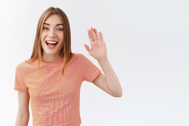 Taillenaufnahme freundliche, hübsche junge Frau in gestreiftem T-Shirt, die Hand hebt und winkt, um Sie zu begrüßen, schön lächelnd, freundlich Hallo sagen, hallo, stehender weißer Hintergrund, der den Kunden im Geschäft begrüßt