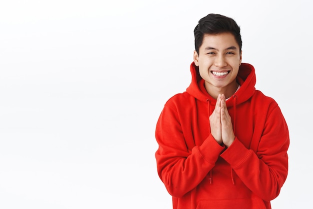Taillenporträt eines glücklichen, fröhlichen asiatischen Hipster-Mannes in rotem Kapuzenpulli, zeigt Namaste-Geste, hält die Handflächen zusammen, um zu beten, lächelnd, dankbar aussehend, erfreut zu beten oder Hilfe zu schätzen