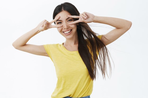 Taillenschuss des erfreuten sorglosen weiblichen Modells im gelben lässigen T-Shirt, das Frieden oder Disco-Geste zeigt