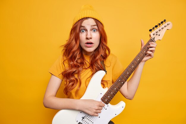 Talentierte Musikerin mit natürlichen roten Haaren sieht schockiert aus, spielt weiße E-Gitarre, trägt Basic-T-Shirt und Hut reagiert auf etwas Überraschendes