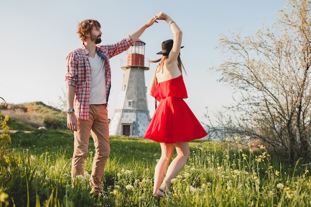 Tanzen junges stilvolles Paar verliebt in Landschaft, Indie-Hipster-Bohème-Stil, Wochenendurlaub, Sommeroutfit, rotes Kleid, grünes Gras, Händchen haltend