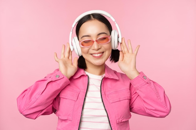Tanzendes stylisches asiatisches Mädchen, das Musik in Kopfhörern hört, die vor rosafarbenem Hintergrund posieren