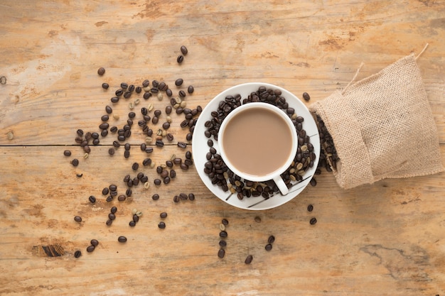 Tasse eines Kaffees mit den gerösteten und rohen Kaffeebohnen, die vom Sack über hölzernem Hintergrund fallen