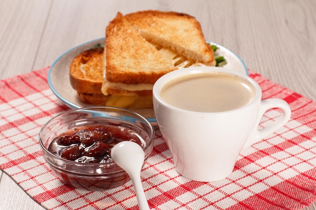 Tasse kaffee, glasschale mit erdbeermarmelade und löffel, geröstete brotscheiben mit käse auf weißem teller mit roter küchenserviette. gutes essen zum frühstück Premium Fotos