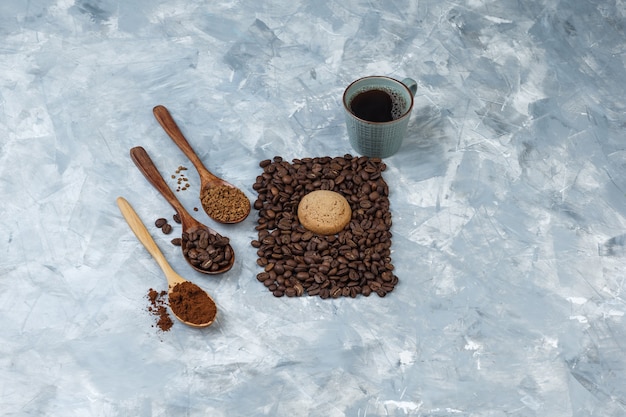 Tasse Kaffee mit hohem Blickwinkel, Keks mit Kaffeebohnen, Instantkaffee, Kaffeemehl in Holzlöffeln auf hellblauem Marmorhintergrund. horizontal