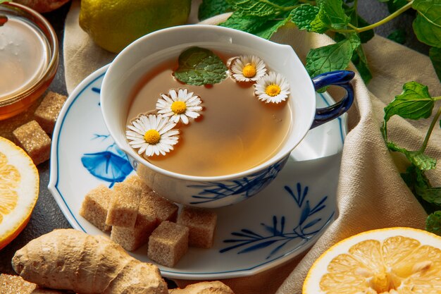 Tasse Kamillentee mit Zitronenscheiben, Ingwer, braunen Zuckerwürfeln und grünen Blättern in einer Untertasse auf grauem und Picknicktuchhintergrund, Nahaufnahme.