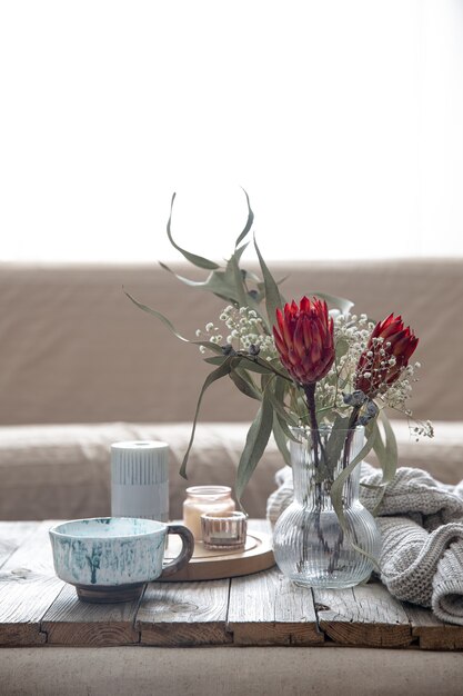 Tasse, Kerzen, Vase mit Protea-Blumen und ein gestricktes Element im Raum auf unscharfem Hintergrund.