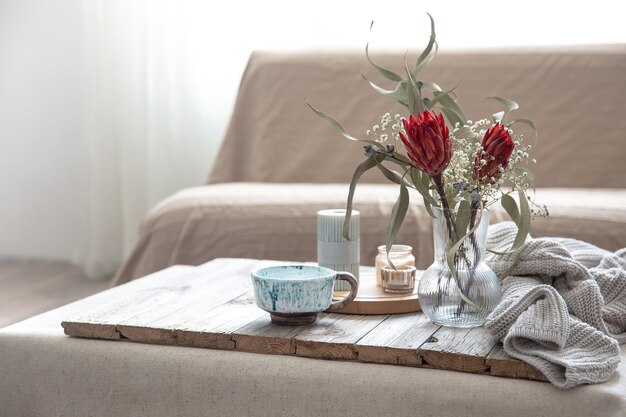 Tasse, Kerzen, Vase mit Protea-Blumen und ein gestricktes Element im Raum auf unscharfem Hintergrund.