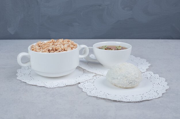 Tasse Kräutertee, Kokosnusskeks und Schüssel mit Süßigkeiten auf weißem Tisch.