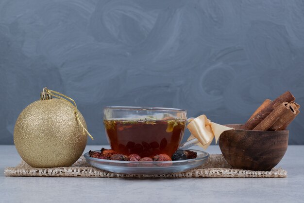 Tasse Tee, Schüssel Zimtstangen und festliche Kugel auf weißem Tisch.