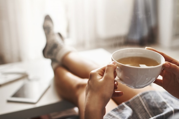 Kostenloses Foto tasse tee und chill. frau, die auf couch liegt, beine auf kaffeetisch hält, heißen kaffee trinkt und morgen genießt, in verträumter und entspannter stimmung ist. mädchen im übergroßen hemd macht pause zu hause
