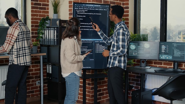Team von Datenbankadministratoren, die den Quellcode auf dem Wandfernseher analysieren und Fehler mit einem digitalen Tablet in einem geschäftigen Serverraum vergleichen. Zwei Cloud-Programmierer debuggen den Algorithmus im Software-Innovationsbüro.
